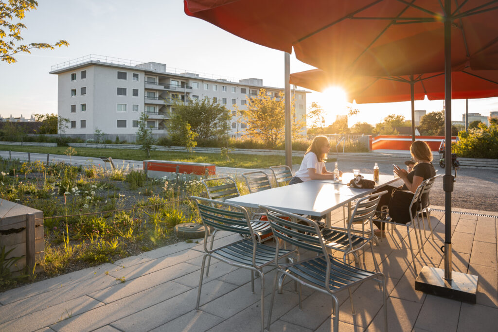 Zwei Frauen sitzen in gemütlicher Abendstimmung in einem Park an einem Gartentisch. Ueberlandpark. Binding Preis für Biodiversität 2026.