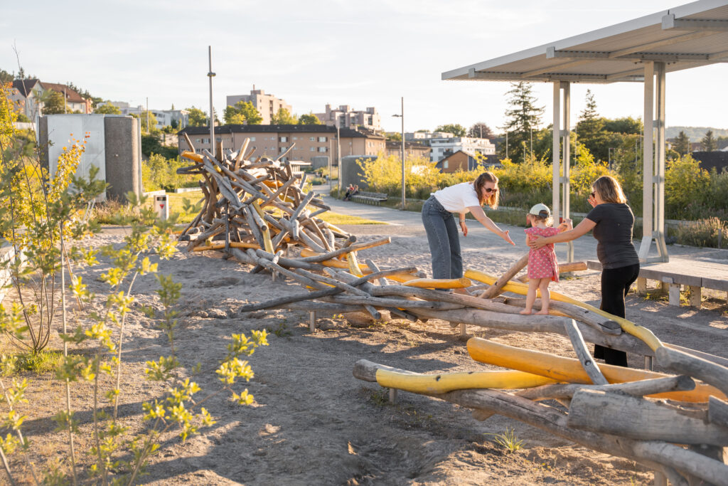 Zwei Frauen spielen mit einem Mädchen auf einem Spielplatz aus Holzstämmen. Binding Preis für Biodiversität 2026.